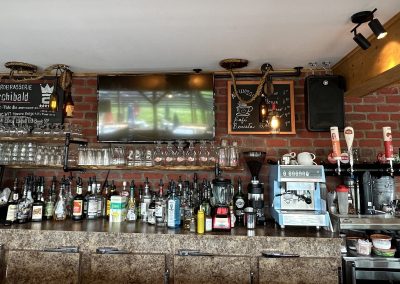 Comptoir d'un bar, avec bouteilles de spiritueux, ardoises et télévision au mur.